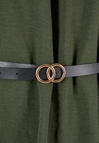 Ceinture en cuir noire avec deux boucles circulaires dorées entrelacées sur un fond en tissu vert foncé.