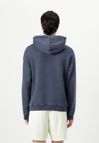 Felpa con cappuccio blu navy, dal taglio rilassato, con una texture morbida e polsini a coste. Presenta un ampio cappuccio e un orlo dritto, abbinata a pantaloni corti chiari.