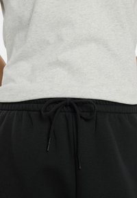 Chemise grise rentrée dans un pantalon de survêtement noir avec un cordon noir noué à la taille élastique, montré au niveau de la taille.