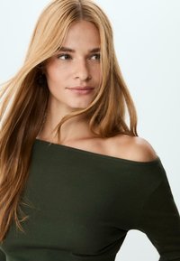 Junge Frau mit langen blonden Haaren, die ein dunkles grünes Off-Shoulder-Oberteil trägt und leicht nach rechts schaut, vor einem schlichten hellen Hintergrund.