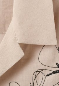 Tissu en lin beige avec un coin plié et une broderie abstraite en fil noir sur une surface texturée.