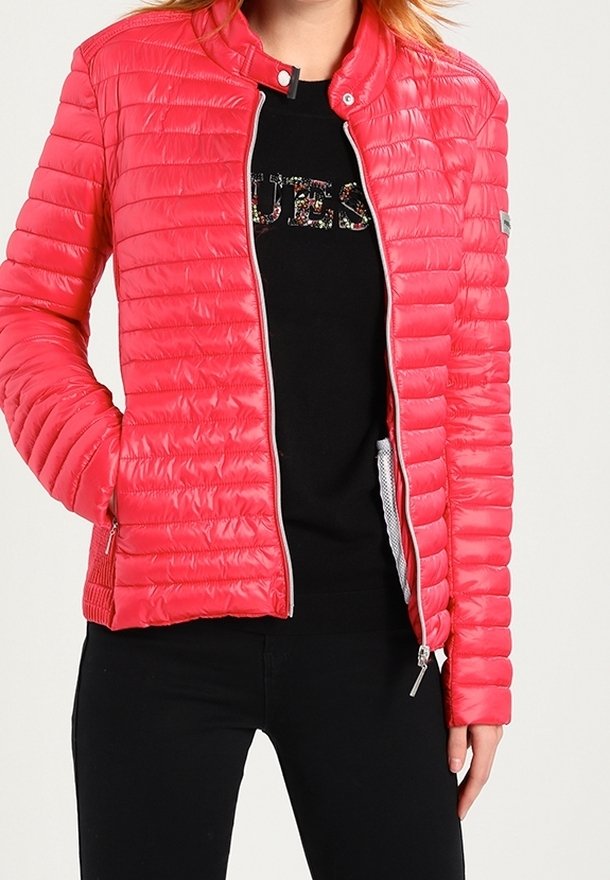 Personne portant une veste matelassée rose vif sur un t-shirt noir avec le texte "GUESS", et un pantalon noir, debout avec une main dans la poche.