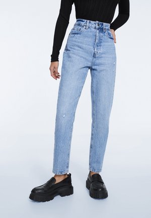 Person iført lyseblå cowboybukser med høj talje og sorte chunky platform loafers, stående med den ene hånd i lommen og den anden hånd afslappet.
