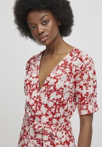 Abito a fiori rosso e bianco con scollo a V profondo, maniche corte e cintura annodata in vita. Tessuto dalla consistenza liscia.