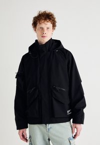Black waterproof jacket with a hood, featuring multiple zippered pockets, adjustable drawstrings, and a relaxed fit. Text detail on the hem.