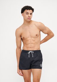 Shorts de bain noirs avec une taille élastique et un cordon blanc. Présente un logo blanc sur la cuisse gauche ; tissu léger et lisse.