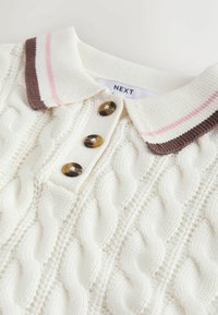 Suéter de punto tipo cable de color crema con cuello a rayas marrón y rosa, y tres botones marrones debajo del cuello.