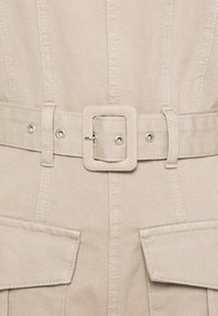 Tissu en denim beige avec une finition texturée, comprenant une ceinture large avec une boucle carrée, et deux poches à rabat à la taille.