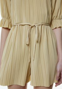Faltenfreies gelbes Jumpsuit mit einer taillierten Taille, kurzen Ärmeln und Rüschensäumen. Weicher, strukturiertes Gewebe mit einer Bindegürtel-Detail.