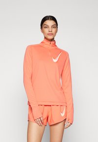 Nike majica dugih rukava u svijetlo koraljnoj boji s polu-zip ovratnikom i bijelim logotipom. Uparen s odgovarajućim koraljnim šortsevima, glatka tkanina, atletski kroj.