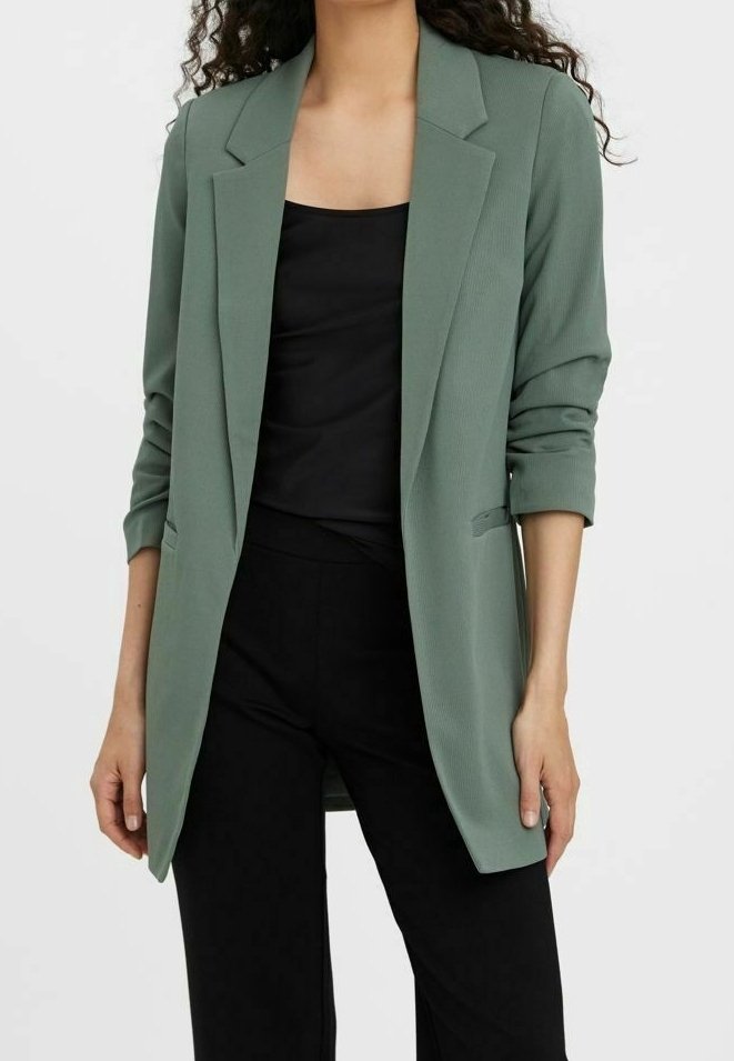 Femme portant un blazer vert clair avec les manches retroussées sur un haut noir et un pantalon noir, debout devant un fonds uni.