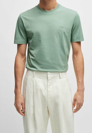 Mann trägt ein hellgrünes kurzärmliges T-Shirt mit „BOSS“-Logo und weiße, hochtaillierte, plissierte Hose vor einfarbigem Hintergrund.