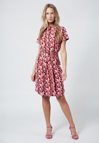 Vestito floreale con maniche corte, vita cintria e orlo fino al ginocchio. Presenta un vivace motivo rosa, rosso e bianco. Indossato con tacchi viola.
