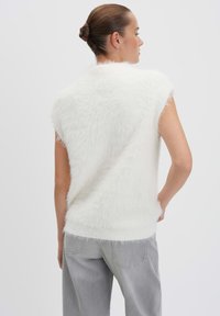 Weißer, ärmelloser Pullover aus flauschigem Material, mit lockerer Passform und geripptem Saum. Getragen mit hellgrauen, leicht baggy geschnittenen Hosen.