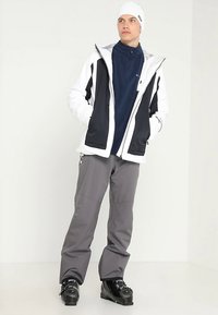 Giacca impermeabile bianca e nera con cappuccio, strato base blu navy con cerniera, pantaloni impermeabili grigi e scarponi da sci neri con dettagli metallici.