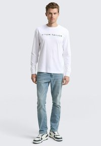 Mann steht da und trägt ein weißes Langarmhemd von Tom Tailor, hellblaue Jeans und weiße Sneakers mit schwarzen Akzenten vor einem einfarbigen Hintergrund.