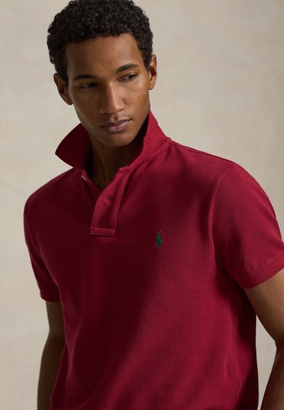 Young man with short curly hair wearing a deep red polo shirt with a raised collar against a plain background.