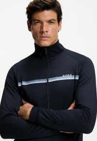 Zwarte sportieve jas met ritssluiting en hoge kraag, met een blauwe en witte horizontale streep en het "BOSS"-logo op de borst. Glad oppervlak.