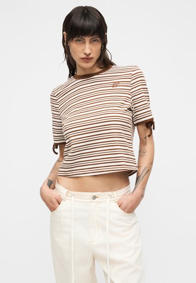 Νεαρή γυναίκα με κοντά σκουρόχρωμα μαλλιά, φορώντας κοντό ριγέ πουκάμισο σε καφέ και άσπρο χρώμα και κρεμ ψηλόμεσα παντελόνια, με τα χέρια στις τσέπες.