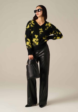 Pull flou noir et jaune avec un décolleté en V, associé à un pantalon en cuir noir taille haute et un sac seau noir texturé. Lunettes de soleil carrées.