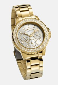 Kulta kello, jossa on hopeanhohtoinen näyttö, kolme alinäyttöä ja kirkkailla jalokivillä koristeltu kehys. Varustettu paksulla metalliketjuhihnalla.