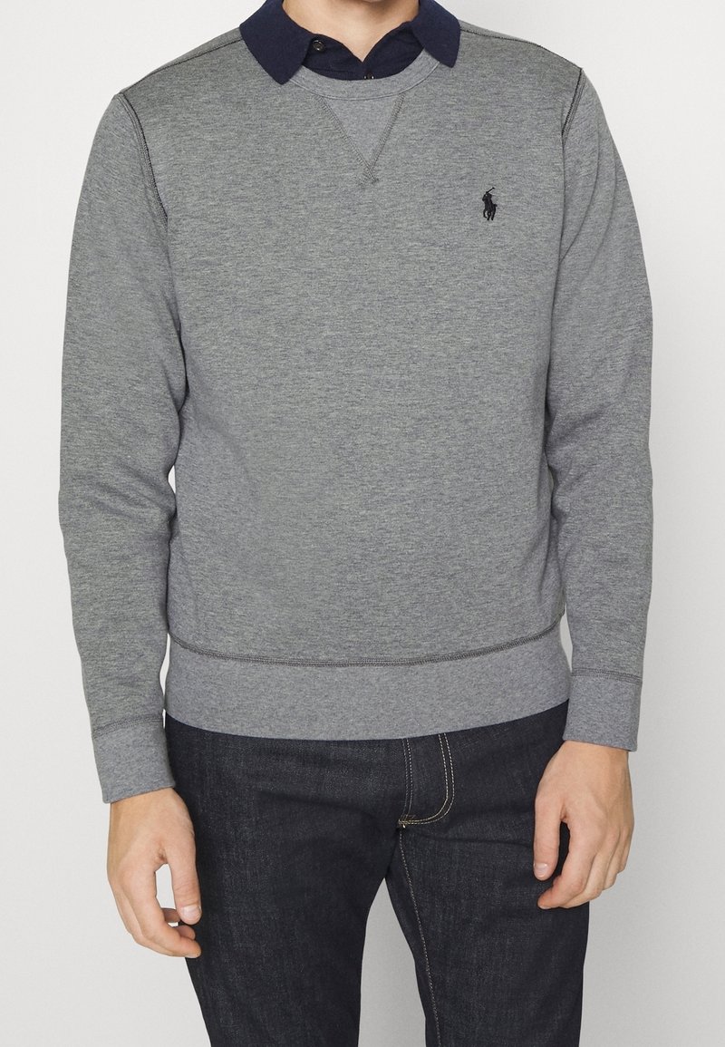 Homme portant un pull gris à manches longues par-dessus une chemise bleu marine à col et un jean foncé, debout devant un fond uni.