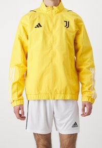 Chaqueta deportiva amarilla con cuello alto, logo negro y tres rayas blancas en cada manga. Ligera, de textura suave y con cierre de cremallera en el frente.