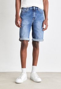 Denimshorts i ljusblått med uppvikta fållar, bärs med vita sneakers och ribbad ankelsocka, vilket visar en avslappnad och ledig stil.