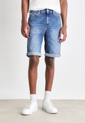 Denimshorts i ljusblått med uppvikta fållar, bärs med vita sneakers och ribbad ankelsocka, vilket visar en avslappnad och ledig stil.