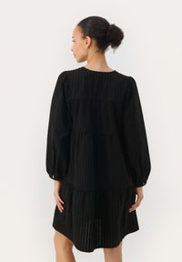 Robe noire à volants avec un tissu texturé, manches bouffantes longues, encolure ronde et ourlet froncé. La coupe est ample.