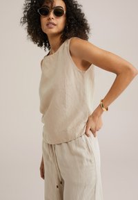 Débardeur beige en lin sans manches avec une coupe décontractée et un col rond, associé à un pantalon ample assorti. Bracelet en or en accent au poignet.