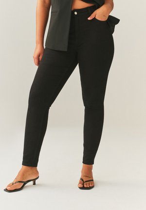 Personne portant un pantalon skinny noir taille haute, avec la main dans la poche et des sandales noires ouvertes à talons kitten sur fond clair.