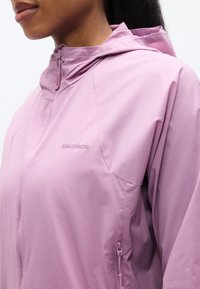 Veste à capuche légère de couleur rose pâle avec une fermeture éclair frontale et des poches latérales. Caractérisée par une texture lisse et un logo discret sur la poitrine.