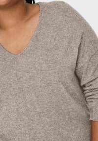 Gros plan d'une personne portant un pull léger à col en V de couleur marron clair, avec des manches longues relevées jusqu'à l'avant-bras.