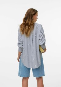 Camicia a righe blu e bianca con fiori ricamati, abbinata a pantaloni corti di jeans azzurri. Maniche lunghe con vestibilità comoda.