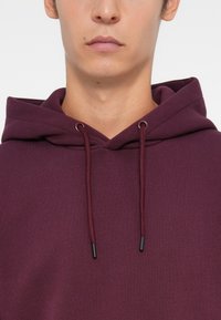 Bordeaux hoodie met een trekkoordkap, voorzien van metaaltippen aan de koorden en een zachte textuur. Het ontwerp heeft een relaxte pasvorm en minimale detailing.