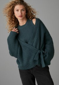 Türkisfarbener gestrickter Pullover mit Wickeldesign und Bindedetail, der ein strukturiertes Muster und weite Ärmel aufweist. Getragen über schwarzen Jeans.