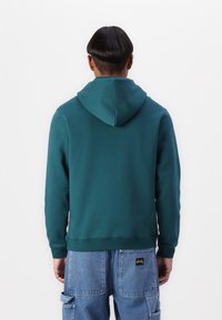 Teal hettegenser med snor, ribbestrikkede ermer og kant. Baksiden har en jevn tekstur, kombinert med blå denim cargo-bukser.