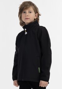 Svart softshelljacka med hög krage, full zip, sidofickor och kontrasterande dragsnören. Tyget verkar slätt och stretchigt.