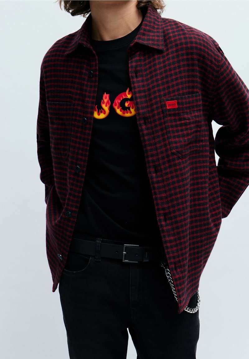 Personne portant une chemise à carreaux rouge et noir boutonnée par-dessus un t-shirt noir avec les lettres en flammes jaunes et rouges "HUGO".