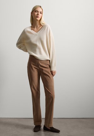 Pull en v crème avec ourlet côtelé, associé à un pantalon en velours côtelé marron et des chaussures à enfiler foncées ; texture lisse et coupe décontractée.