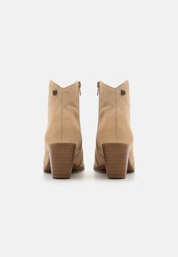 Botas de tobillo de ante beige con un ligero tacón, que presentan una cremallera en la parte trasera y un distintivo cuello en forma de V. Textura suave, con acentos de hardware minimalistas.