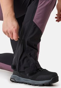 Pantaloni outdoor neri impermeabili con accenti viola, gambe inferiori con zip e chiusura a strappo. Indossati con scarpe da trekking nere.