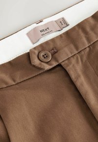 Brown relaxed fit trousers featuring a smooth texture, top button closure, and white inner band, with a tag displaying size and brand information.