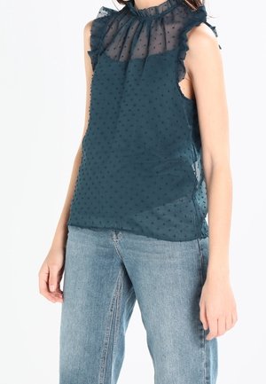 Femme portant un chemisier transparent sans manches vert sarcelle foncé avec des volants et une texture à pois, associé à un jean bleu clair.