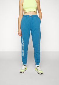 Blue sweatpants with a drawstring waist, featuring a white "Quiksilver" logo on the left leg. Worn with a bright lime green crop top and sneakers.