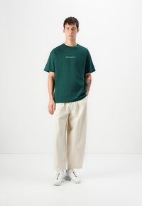 Grønn bomull t-skjorte med korte ermer og rund hals, med hvit tekst på forsiden. Kombinert med beige løst sittende bukser og hvite joggesko.