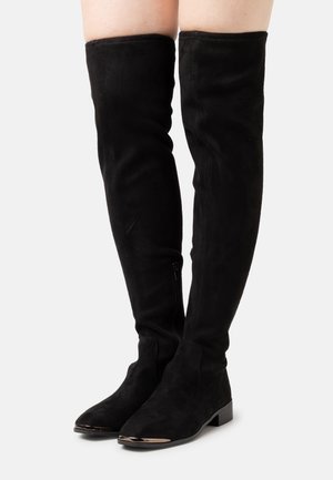 Bottes hautes en daim noir dépassant le genou, avec talons bas et fermetures éclair latérales, portées sur des jambes sur un fond blanc uni.