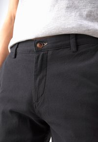 Svarta byxor i texturerad tyg, med en knappstängning på framsidan med en brun knapp och fickdetalj, visade från midjan och ner.