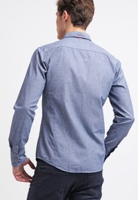 Blue patterned long-sleeve shirt made of fabric with a smooth texture. Features a buttoned cuff and a curved hemline. Note: plain back view.